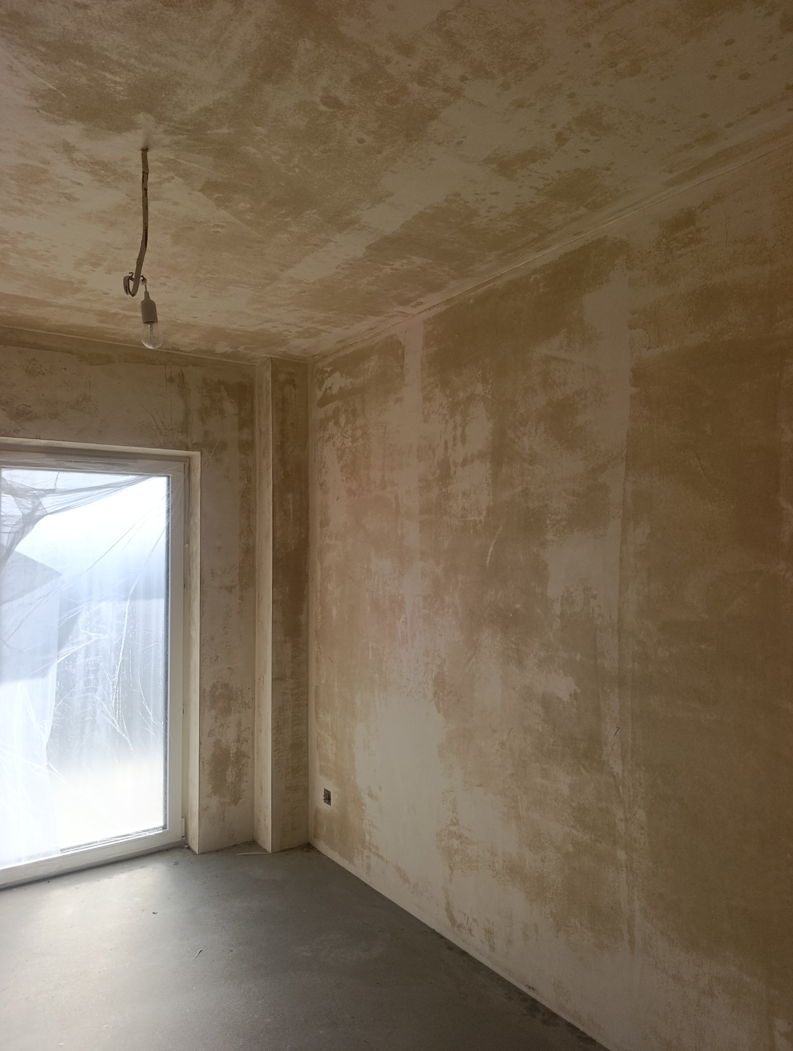 Pokój w trakcie przygotowania do malowania: surowe ściany z widocznymi śladami szpachlowania, okno zabezpieczone folią, sufit z wiszącą żarówką. Stan deweloperski.