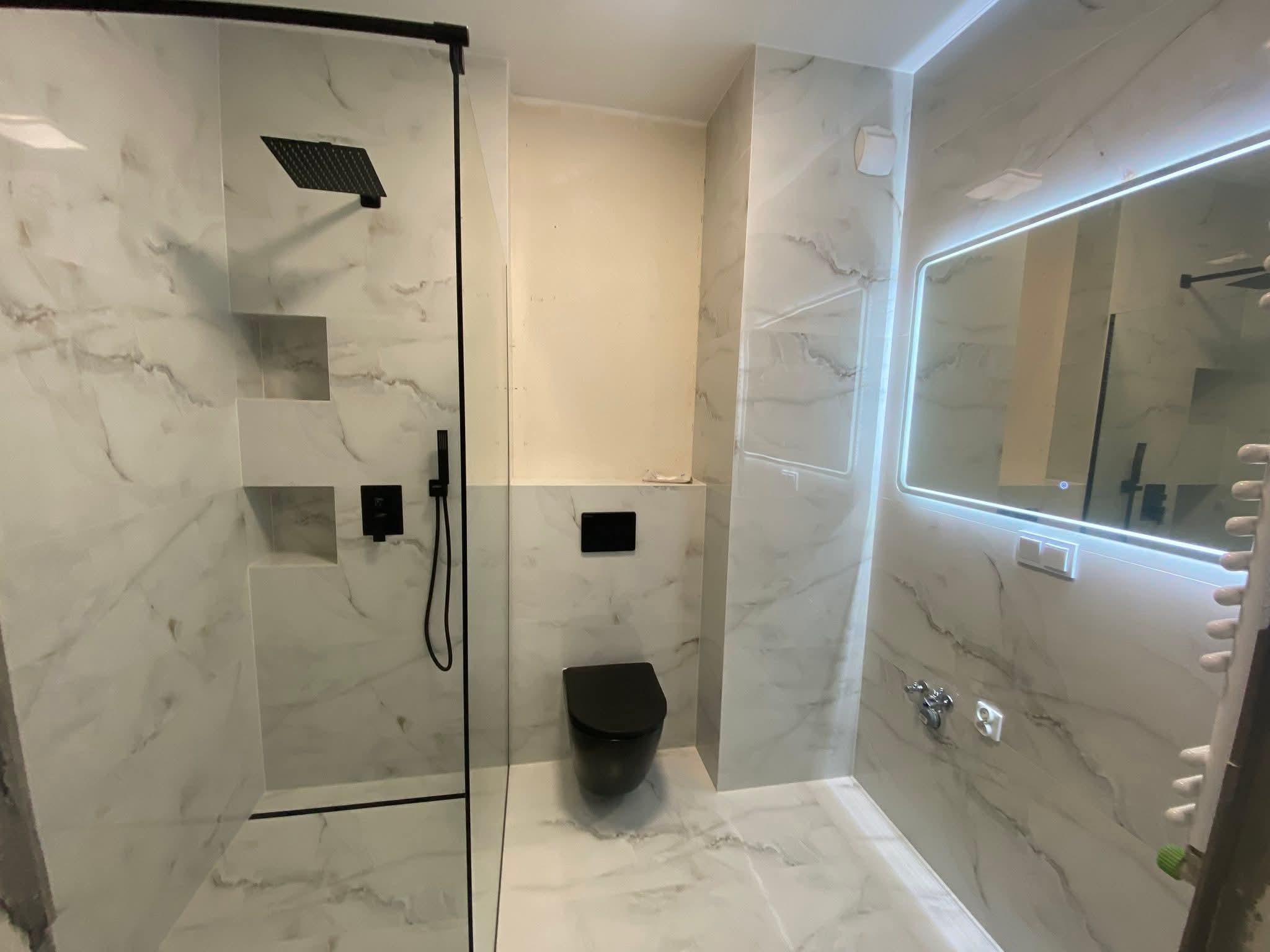 Nowoczesna łazienka z kabiną typu walk-in, wykończona białym marmurem, czarną armaturą i podświetlanym lustrem. Minimalistyczny design z akcentami czerni.