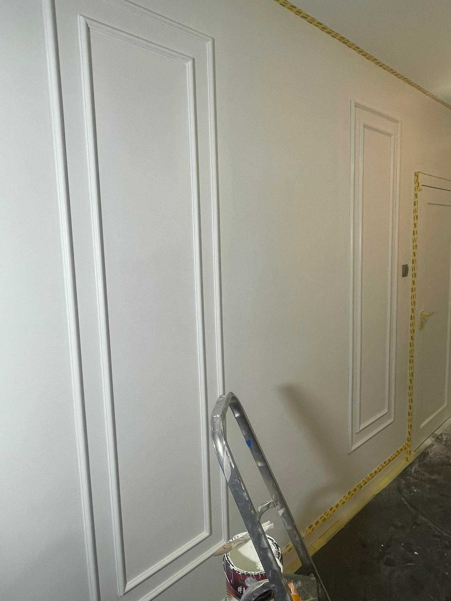 Ściana z dekoracyjnymi panelami pomalowana na jasny odcień szarości, zabezpieczona taśmą malarską, z widoczną drabiną i puszką farby w dolnym rogu.