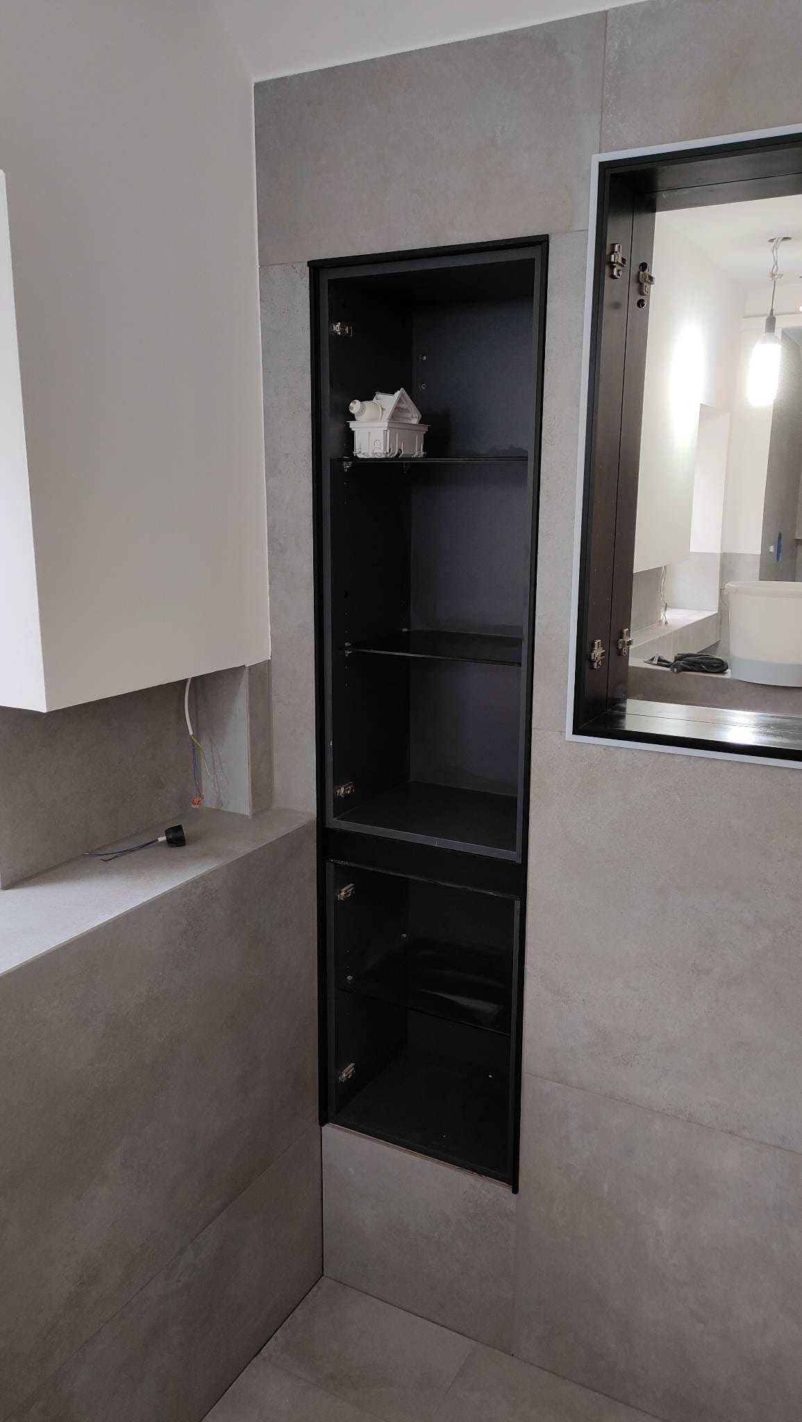 Wnęka z czarną szafką w szarej łazience, z otwartymi drzwiczkami i dekoracyjnym domkiem na półce. Minimalistyczny design, płytki imitujące beton.