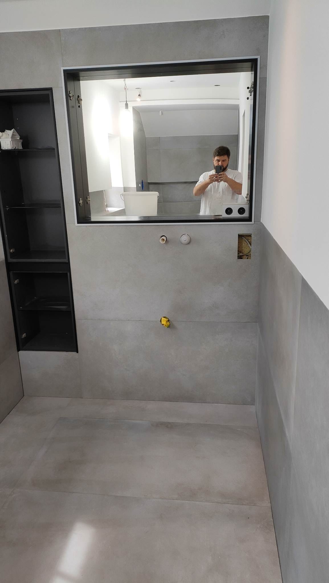 Szara łazienka w trakcie wykańczania: ułożone płytki, zamontowane lustro z odbiciem osoby robiącej zdjęcie, widoczna zabudowa meblowa i instalacje. Minimalistyczny styl.