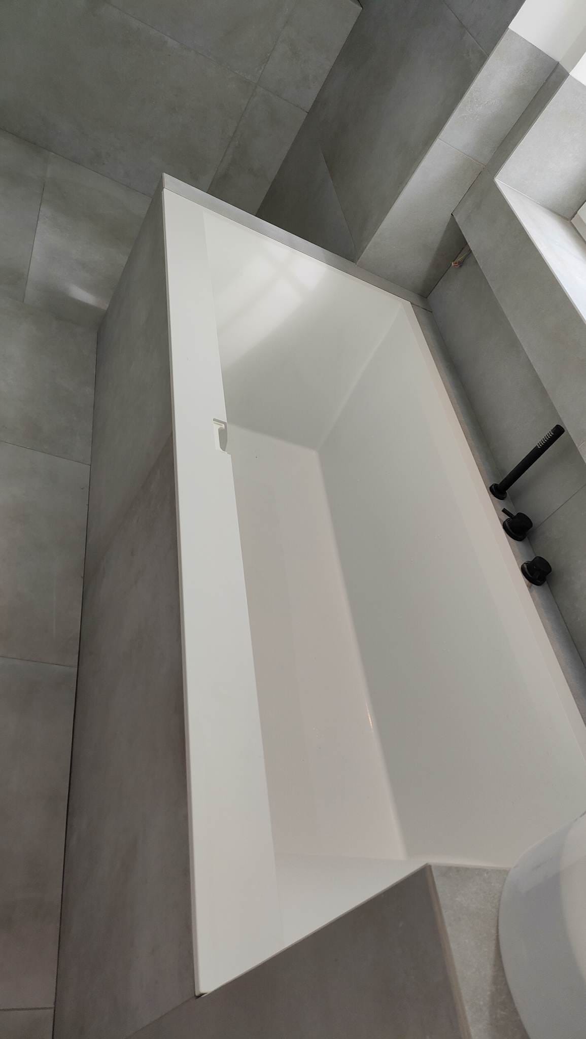Nowoczesna, biała wanna w minimalistycznej łazience wykończonej szarymi płytkami. Widoczna czarna bateria i fragment umywalki. Ujęcie z góry, podkreślające geometrię.