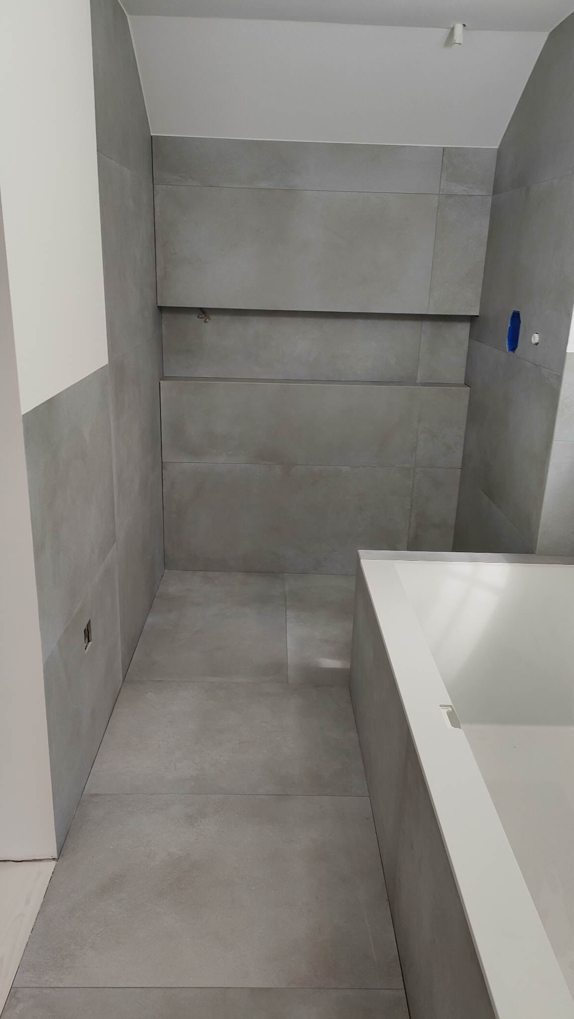 Minimalistyczna łazienka w szarościach: duże, matowe płytki na ścianach i podłodze, zabudowana wanna z białym obramowaniem. Nowoczesne wykończenie wnętrza.