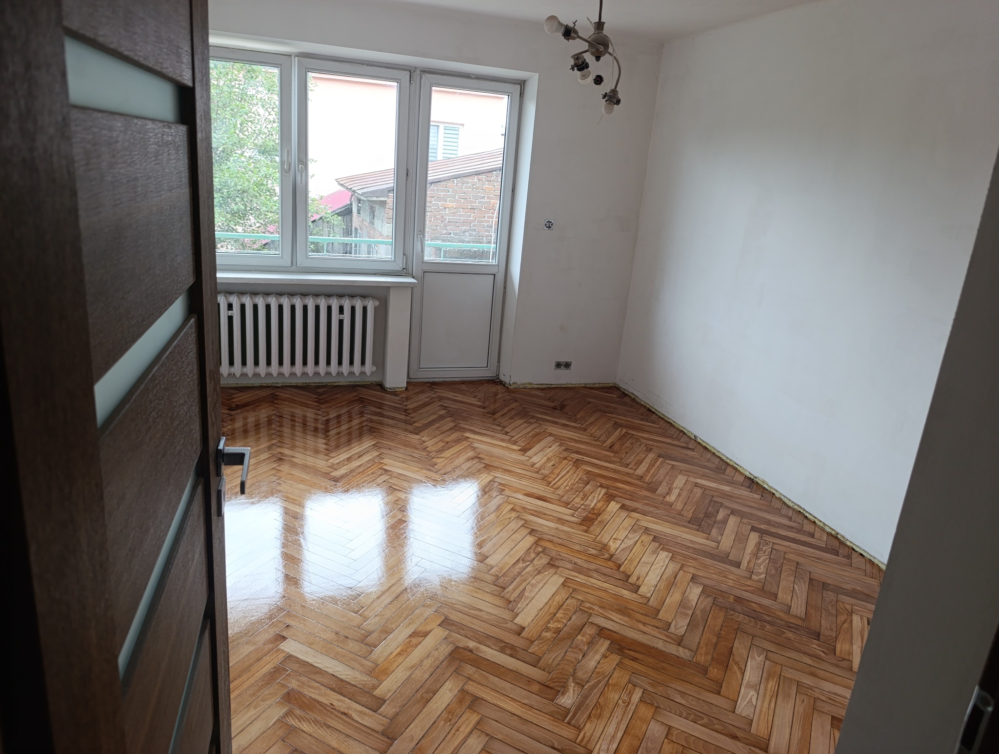 Pokój z parkietem ułożonym w jodełkę, widok zza uchylonych drzwi. Świeżo położona podłoga odbija światło z okna. Białe ściany i grzejnik.