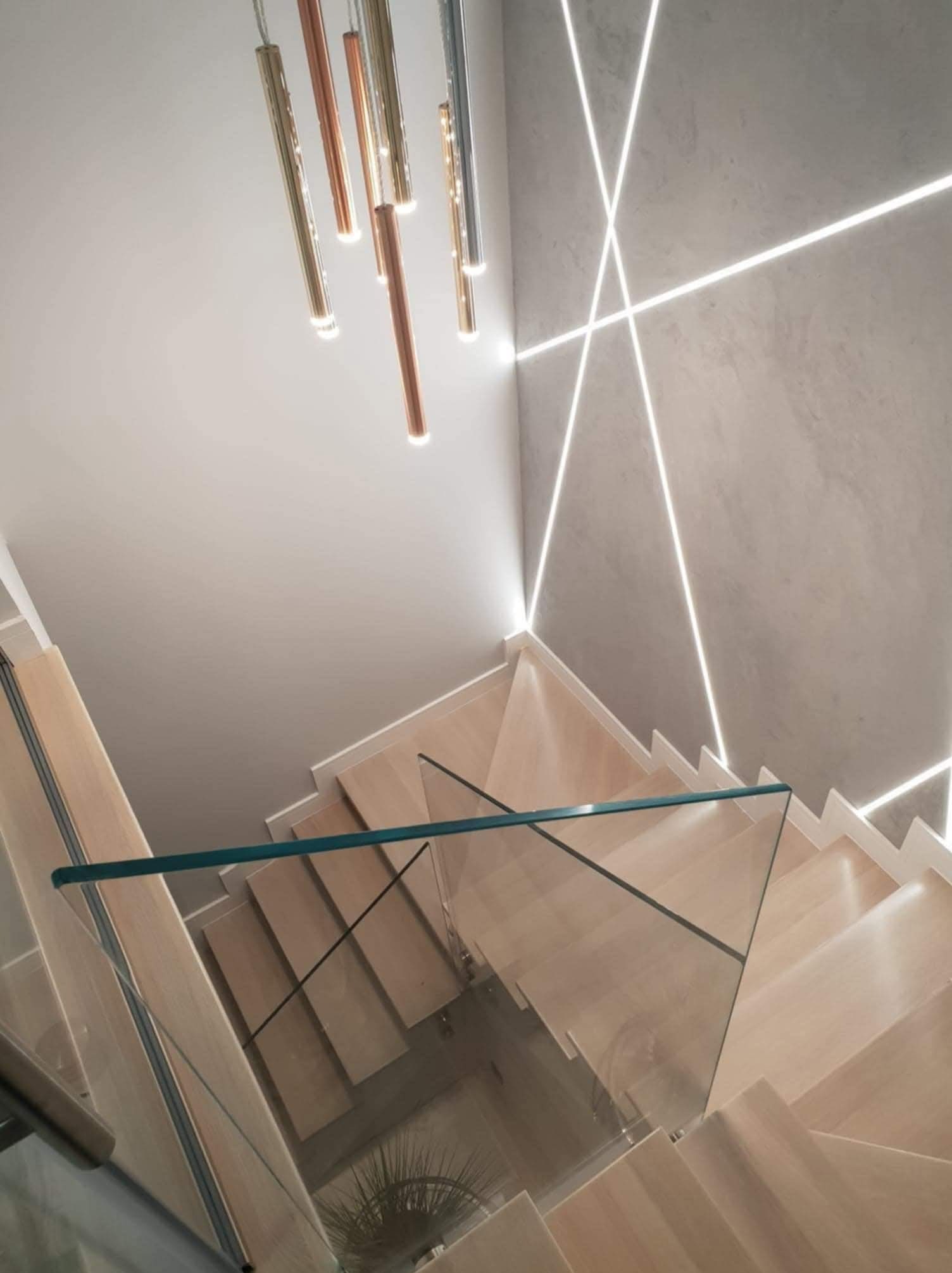 Nowoczesne schody z drewnianymi stopniami, szklaną balustradą i dekoracyjnym oświetleniem LED na ścianie oraz wiszącą lampą z metalowych rurek.