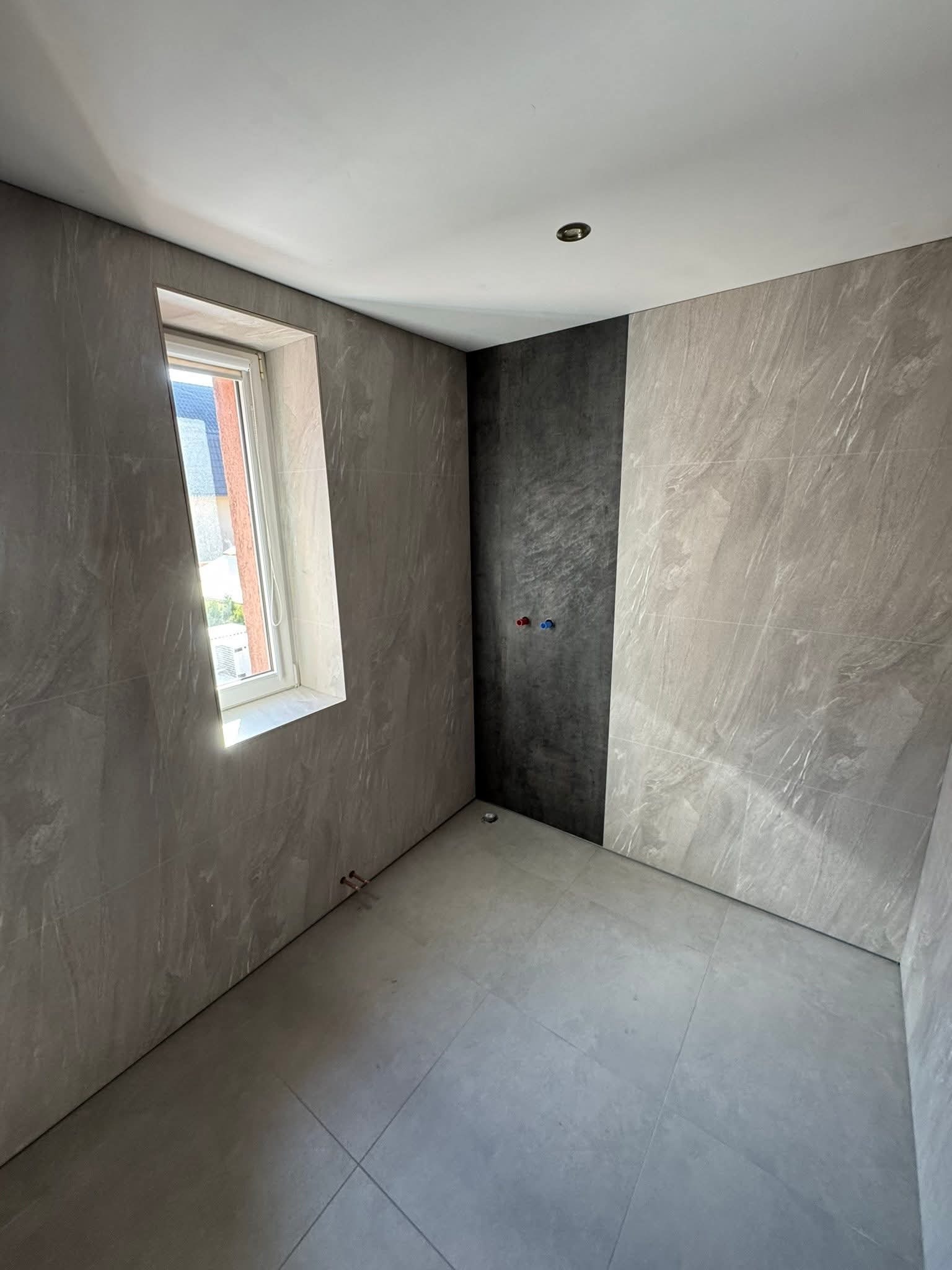 Wykończona łazienka z szarymi płytkami imitującymi kamień, z oknem i fragmentem czarnej ściany. Widoczne przyłącza wodne. Minimalistyczny design.