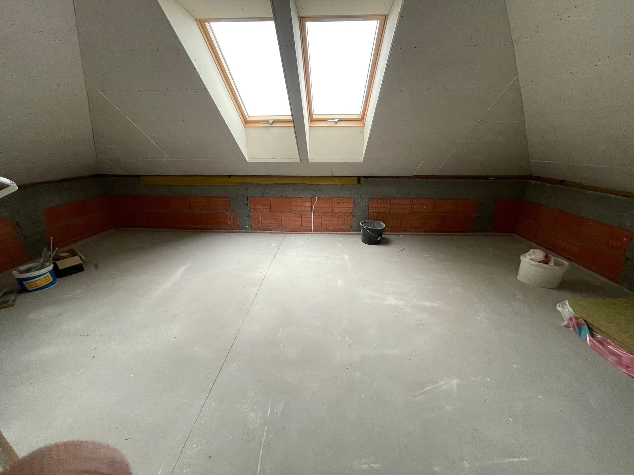Poddasze w trakcie adaptacji: surowe ściany z cegły, okna dachowe, szpachlowana podłoga. Widoczne narzędzia i materiały budowlane. Wnętrze w stanie przed wykończeniem.