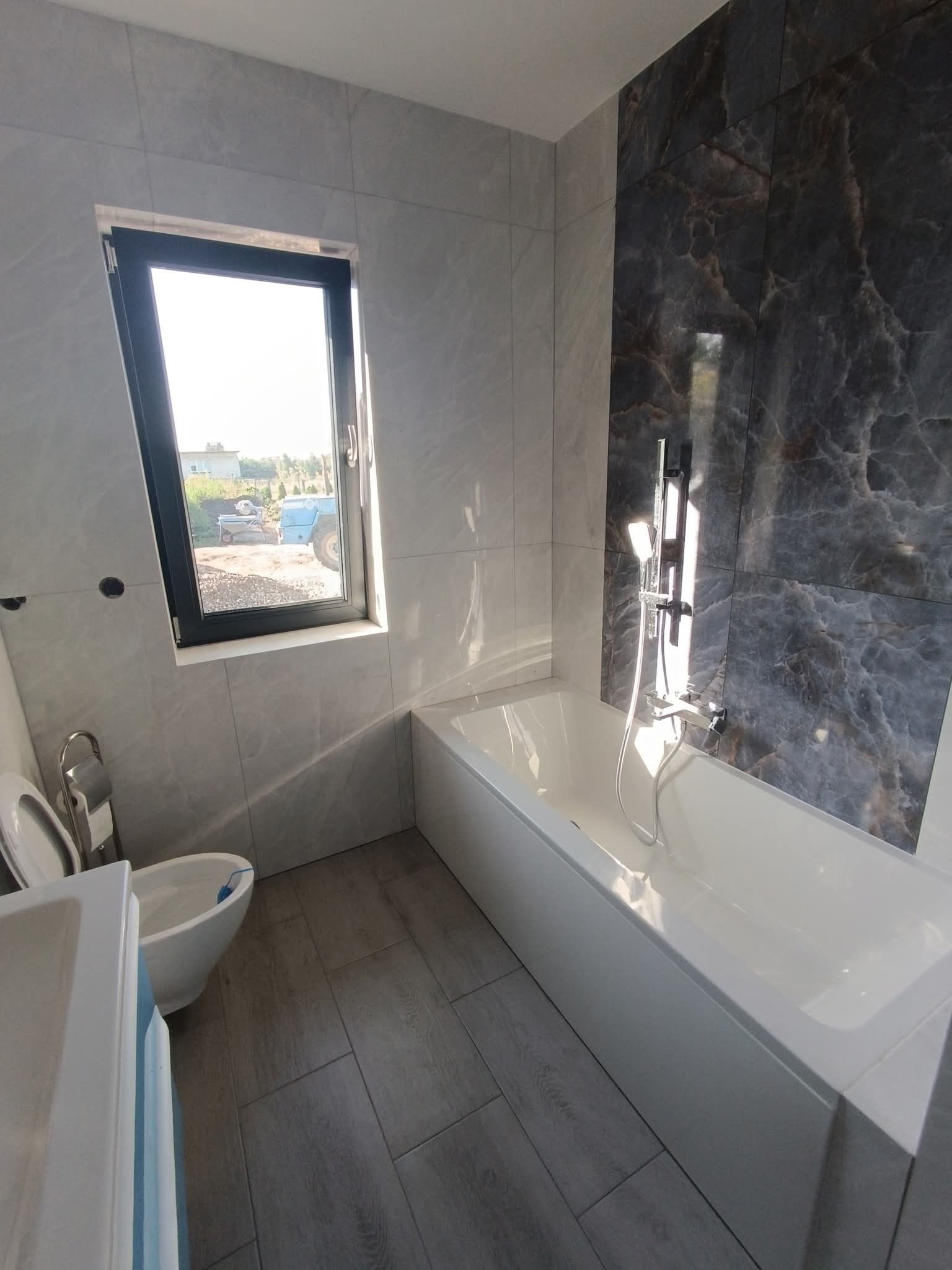 Nowoczesna łazienka z marmurową ścianą, białą wanną i bidetem. Okno z widokiem na zewnątrz. Szare płytki podłogowe imitujące drewno. Minimalistyczny design.