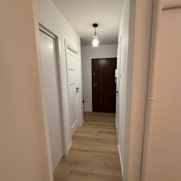 M.Art - Jasny korytarz z drewnopodobną podłogą i białymi drzwiami. W tle widoczna ciemna, drewniana brama wejściowa i designerska lampa sufitowa. Minimalistyczny wystrój.