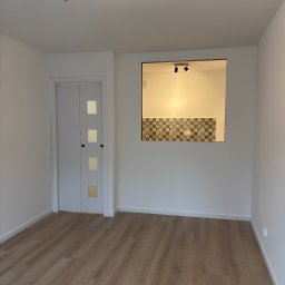 M.Art - Jasne wnętrze po odświeżeniu: drewniana podłoga, białe ściany, drzwi z przeszkleniami i widok na fragment kuchni z dekoracyjnymi płytkami. Nowoczesne oświetlenie sufitowe.