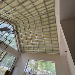 M.Art - Montaż izolacji stropu z wełny mineralnej w metalowym stelażu. Widoczne okna i fragment rusztowania. Prace wykończeniowe w jasnym wnętrzu.