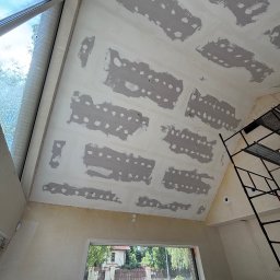 M.Art - Sufit poddasza w trakcie prac: płyty gipsowe z widocznymi łączeniami szpachlowanymi, okno dachowe i rusztowanie. Widok na zewnątrz przez okno.