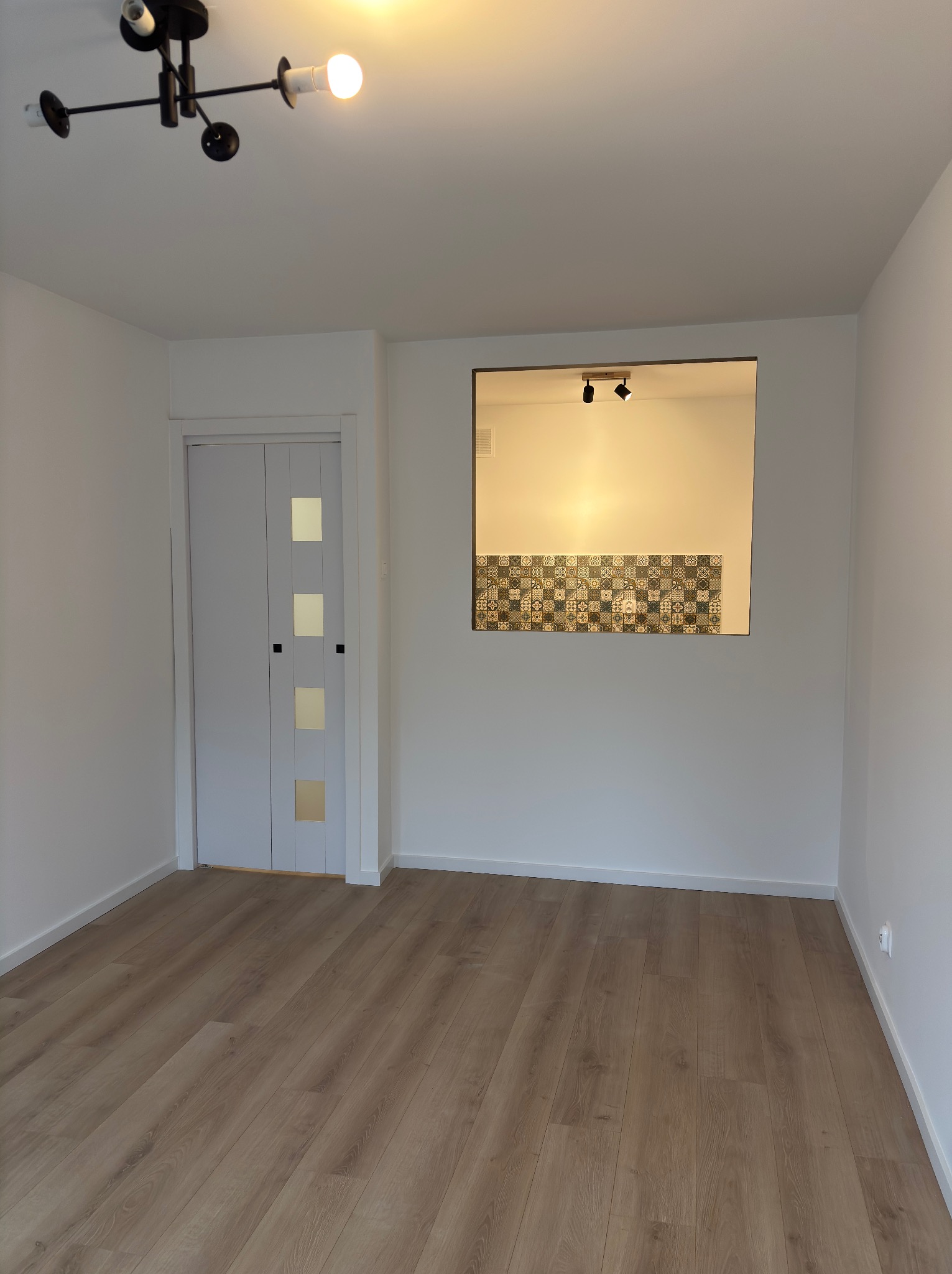 Jasne wnętrze po odświeżeniu: drewniana podłoga, białe ściany, drzwi z przeszkleniami i widok na fragment kuchni z dekoracyjnymi płytkami. Nowoczesne oświetlenie sufitowe.