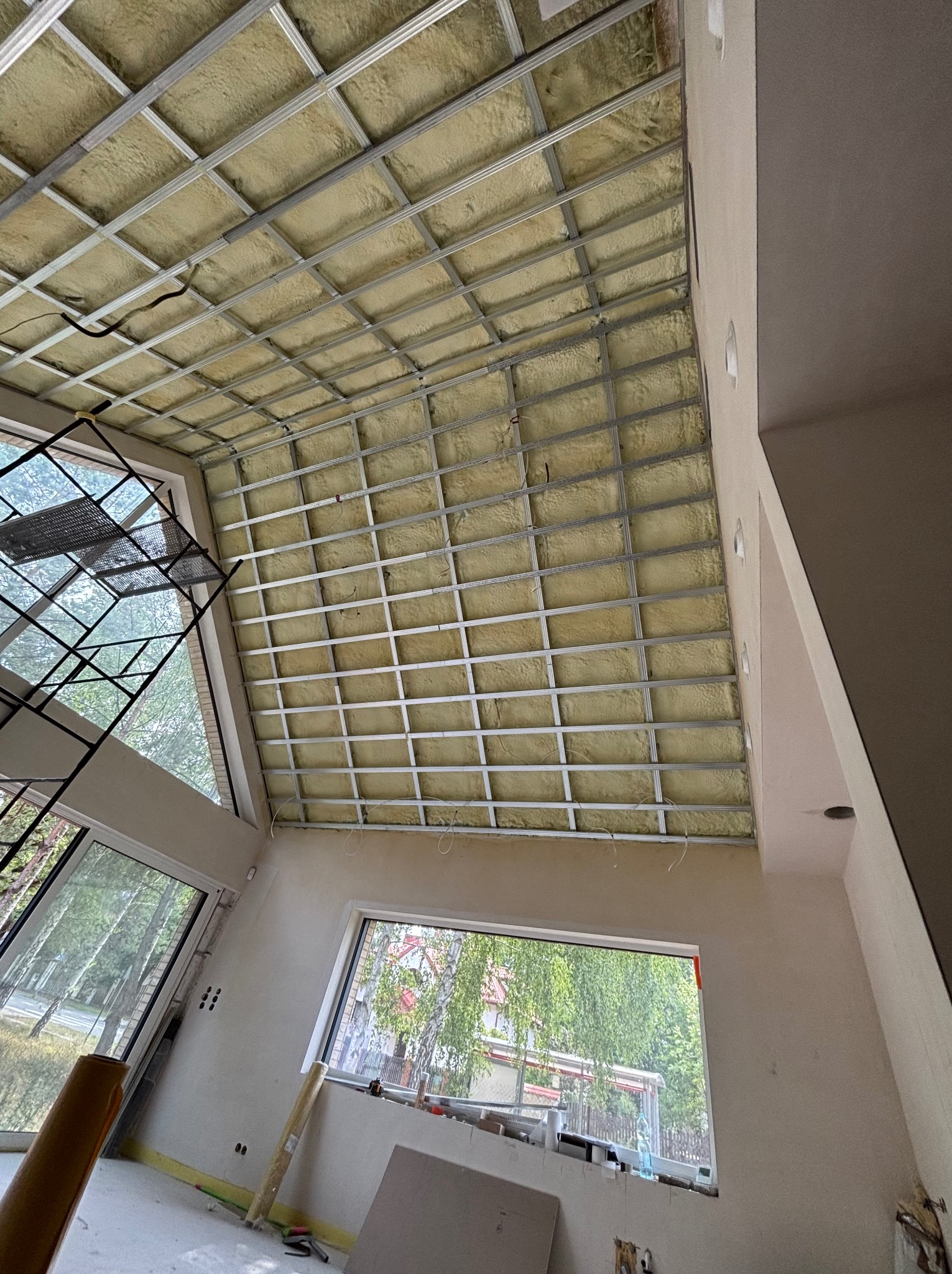 Montaż izolacji stropu z wełny mineralnej w metalowym stelażu. Widoczne okna i fragment rusztowania. Prace wykończeniowe w jasnym wnętrzu.