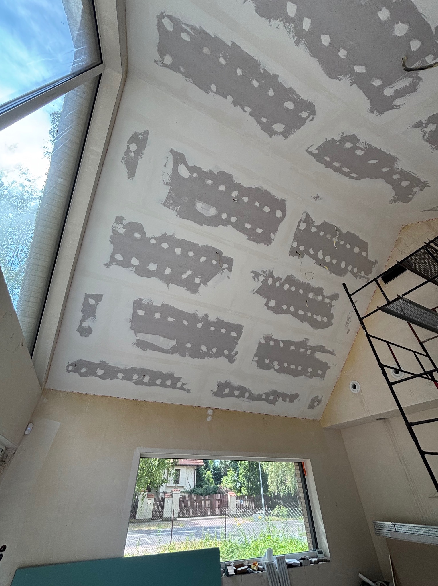 Sufit poddasza w trakcie prac: płyty gipsowe z widocznymi łączeniami szpachlowanymi, okno dachowe i rusztowanie. Widok na zewnątrz przez okno.