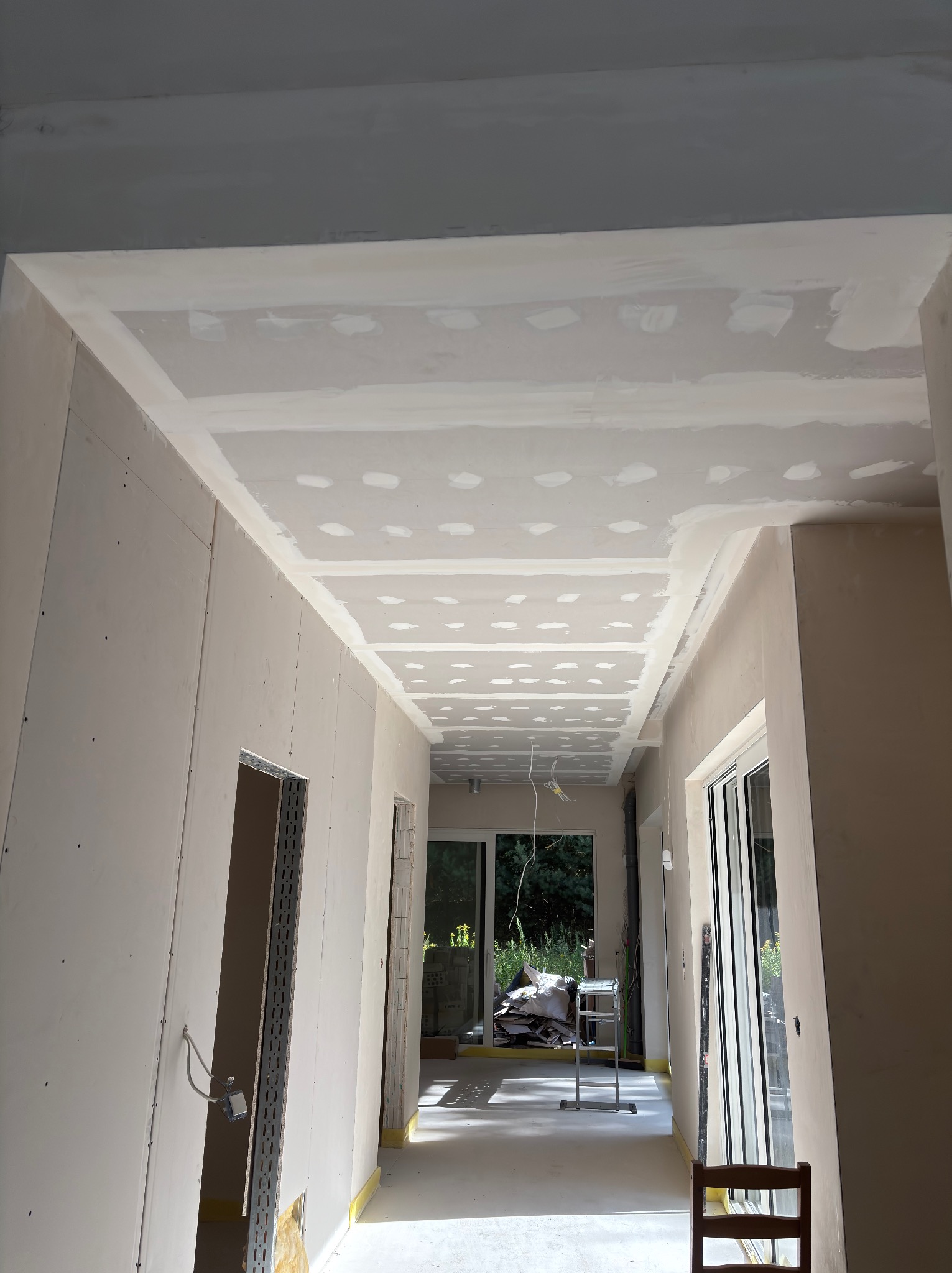 Korytarz w trakcie prac wykończeniowych: ściany i sufit pokryte płytami gipsowo-kartonowymi, widoczne łączenia wymagające szpachlowania. W tle okna i elementy rusztowania.
