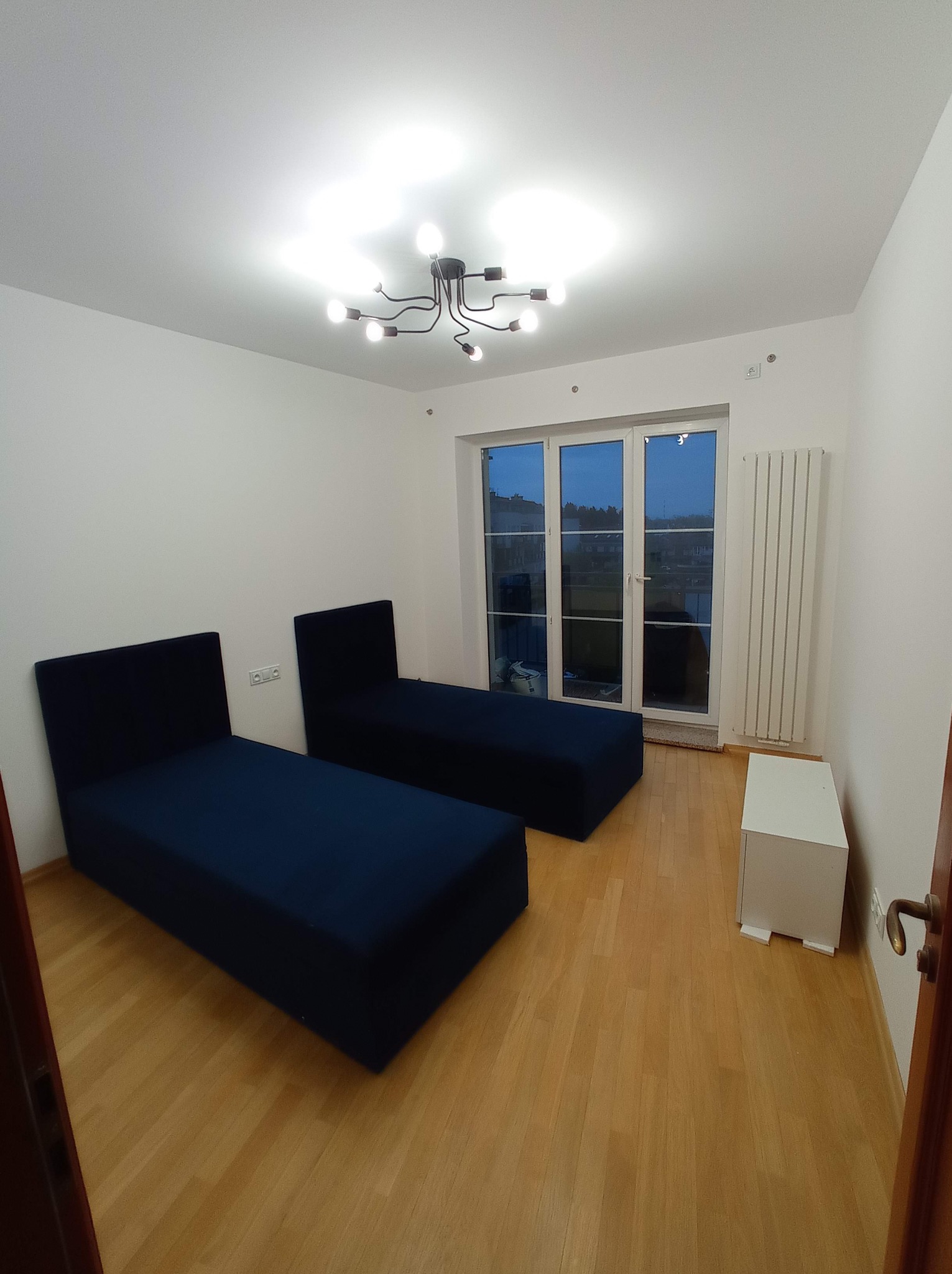 Jasne wnętrze pokoju z dwoma granatowymi tapczanami, drewnianą podłogą i białym grzejnikiem pod oknem. Nowoczesna lampa sufitowa.