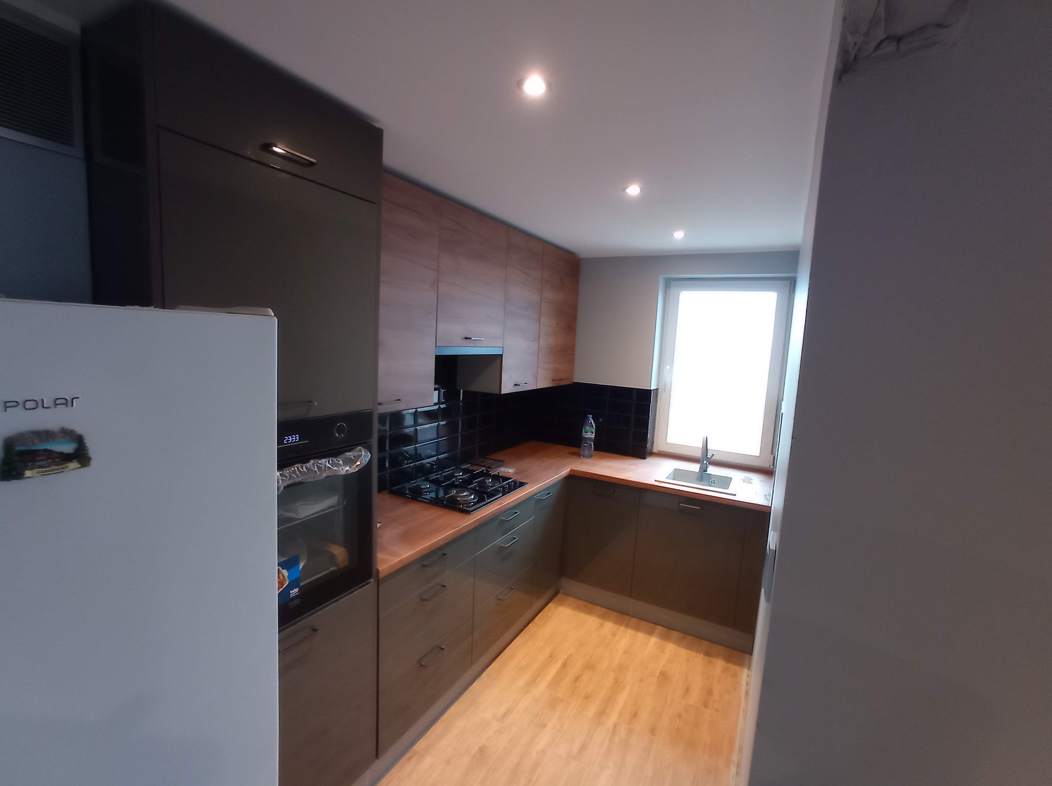 Nowoczesna kuchnia z drewnianymi blatami i czarnymi płytkami nad kuchenką. Widoczny fragment lodówki i okno zlewem. Uwagę przykuwa minimalistyczny design i oświetlenie.