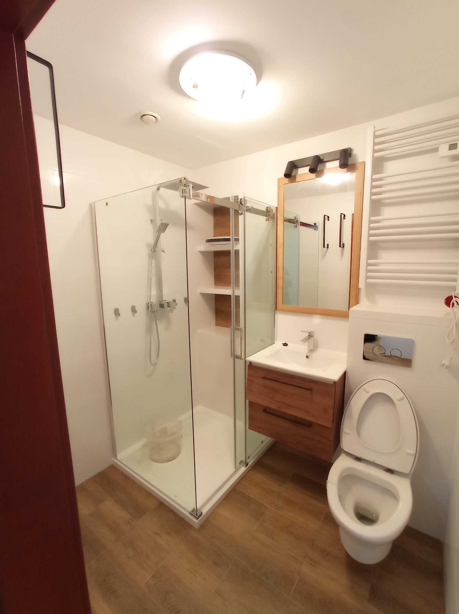 Nowoczesna łazienka po remoncie: kabina prysznicowa ze szklanymi drzwiami przesuwnymi, drewniana szafka pod umywalką, toaleta i grzejnik drabinkowy.