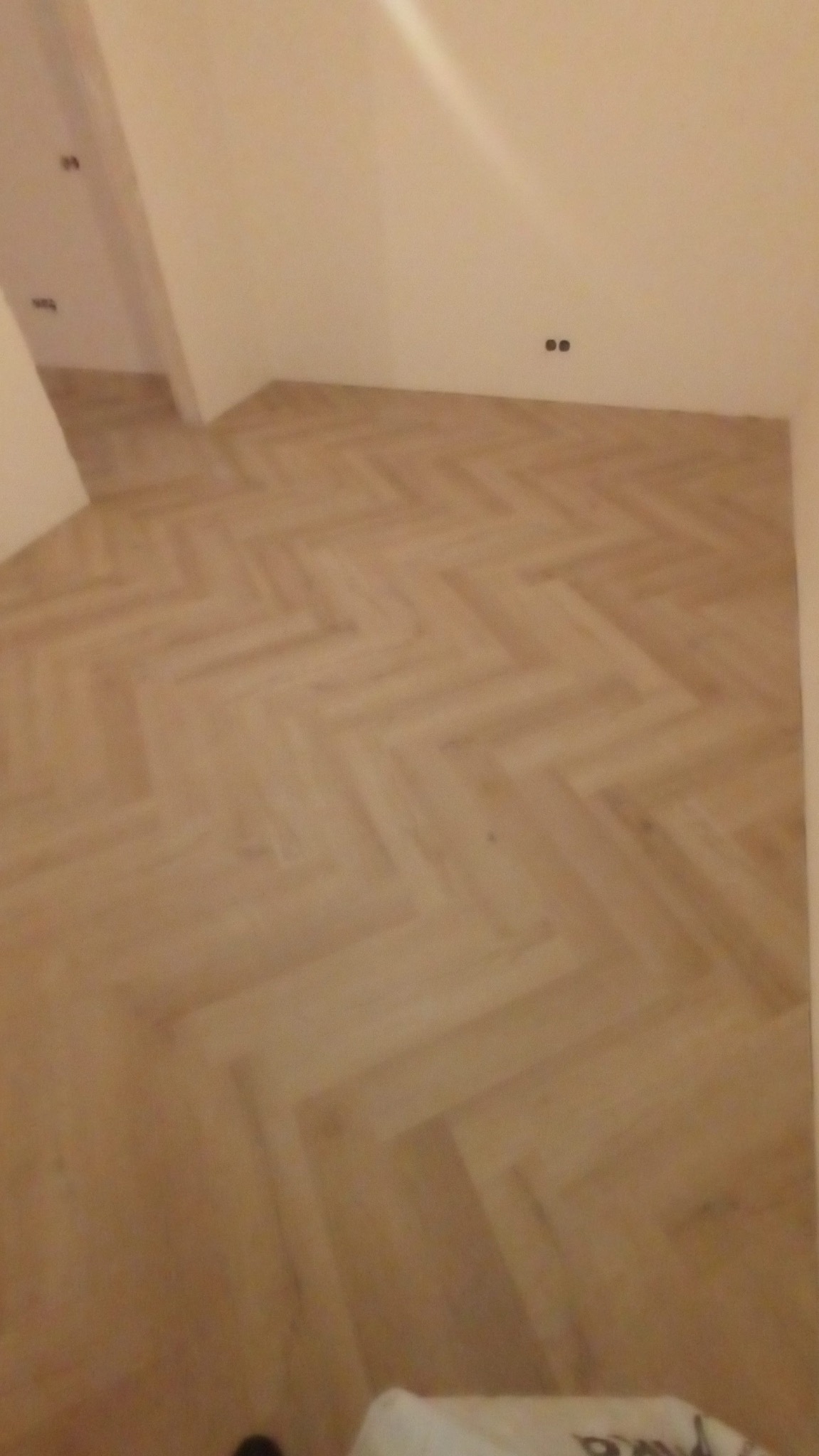 Jasna, drewniana podłoga ułożona w jodełkę w pustym pokoju z białymi ścianami. Widoczne gniazdka elektryczne na ścianie i fragment progu w przejściu do innego pomieszczenia.