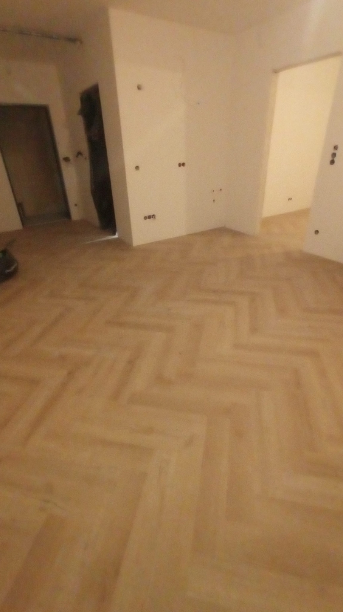 Ukończona podłoga z parkietu ułożonego w jodełkę w pustym, odświeżonym pokoju z białymi ścianami i widocznymi otworami na gniazdka elektryczne.