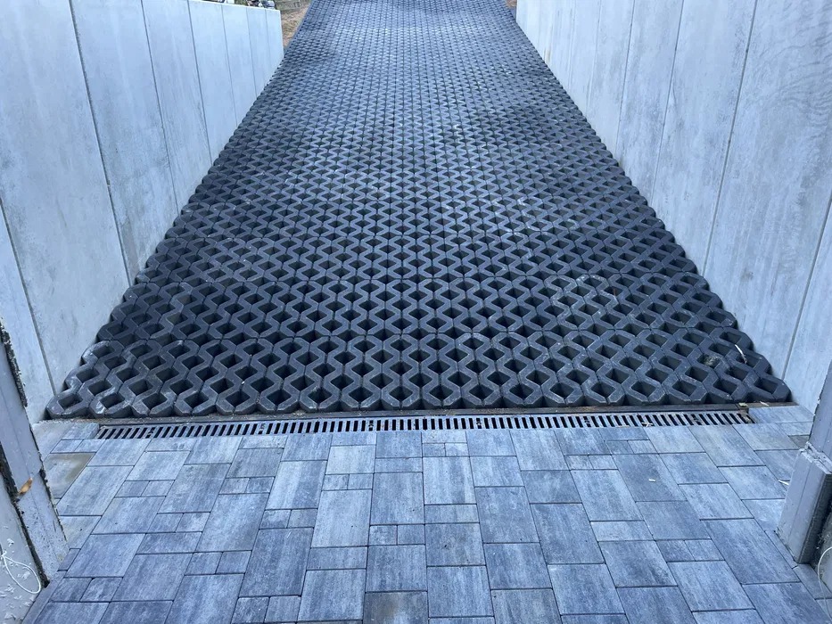 Ukończona nawierzchnia z ażurowych płyt betonowych na pochyłym terenie, oddzielona od kostki brukowej linią odpływu liniowego, otoczona betonowymi ścianami.