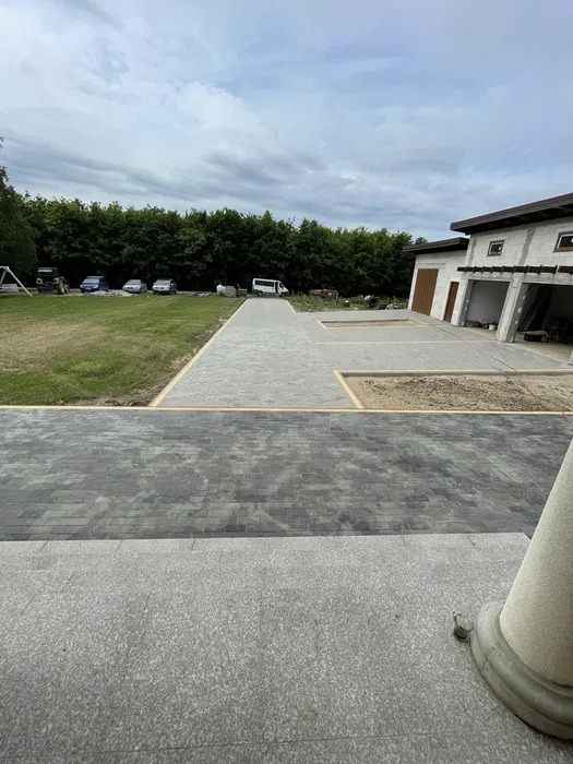 Ułożona kostka brukowa w dwóch odcieniach szarości prowadząca do domu z garażem, widok z góry, w tle trawnik i drzewa. Częściowo widoczny filar.