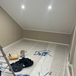 Łapka Remontuje - Pokój na poddaszu w trakcie malowania ścian na beżowo, zabezpieczony folią malarską. Widoczna drabina, puszka z farbą i narzędzia malarskie.