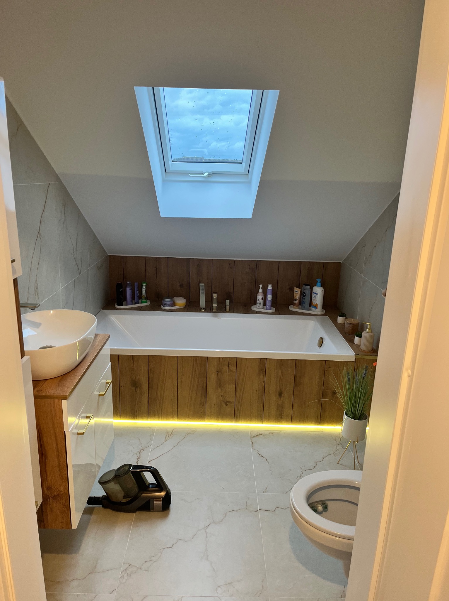 Nowoczesna łazienka na poddaszu z wanną obudowaną drewnem, oświetleniem LED, umywalką nablatową i marmurową podłogą. Widok zza uchylonych drzwi.