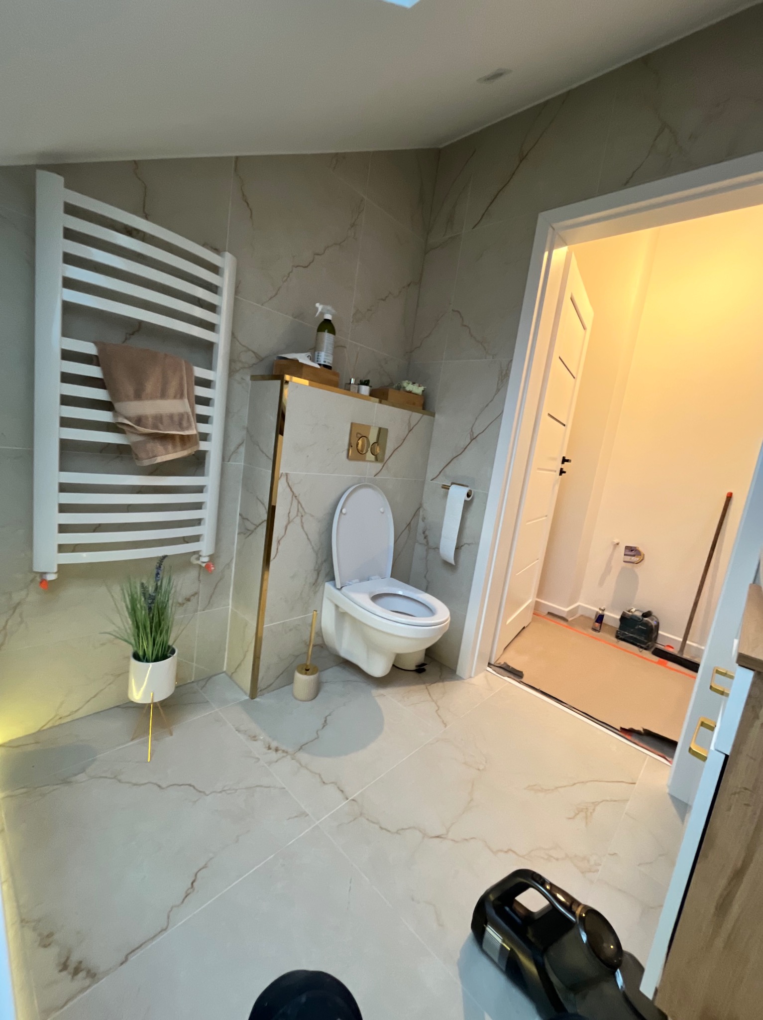 Jasna łazienka z marmurowymi płytkami, grzejnikiem drabinkowym, toaletą i złotymi akcentami. Widoczny fragment odkurzacza.