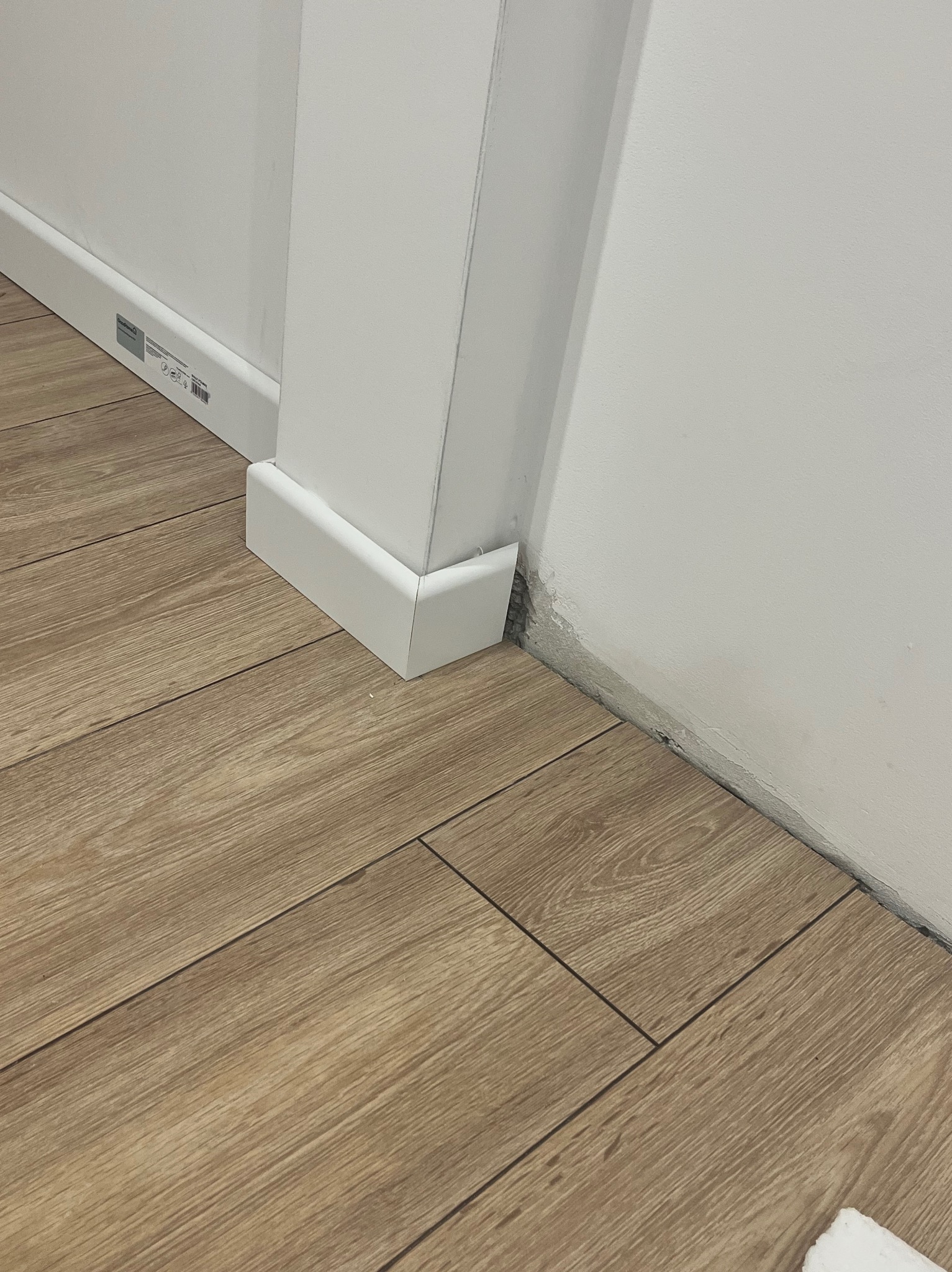 Ułożona podłoga z paneli imitujących drewno, biała listwa przypodłogowa i fragment ościeżnicy. Widoczne niedociągnięcia w wykończeniu ściany przy podłodze.