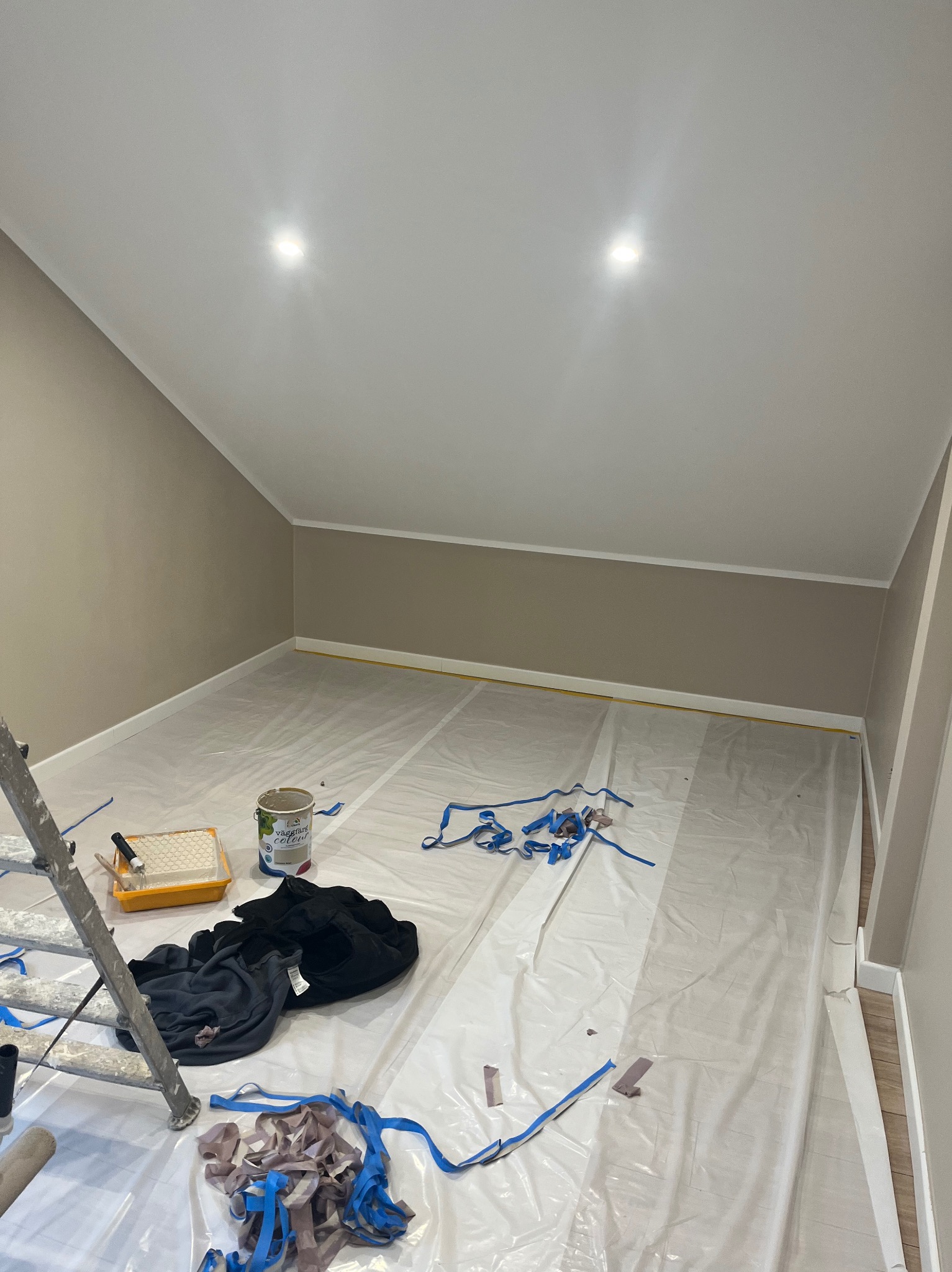 Pokój na poddaszu w trakcie malowania ścian na beżowo, zabezpieczony folią malarską. Widoczna drabina, puszka z farbą i narzędzia malarskie.