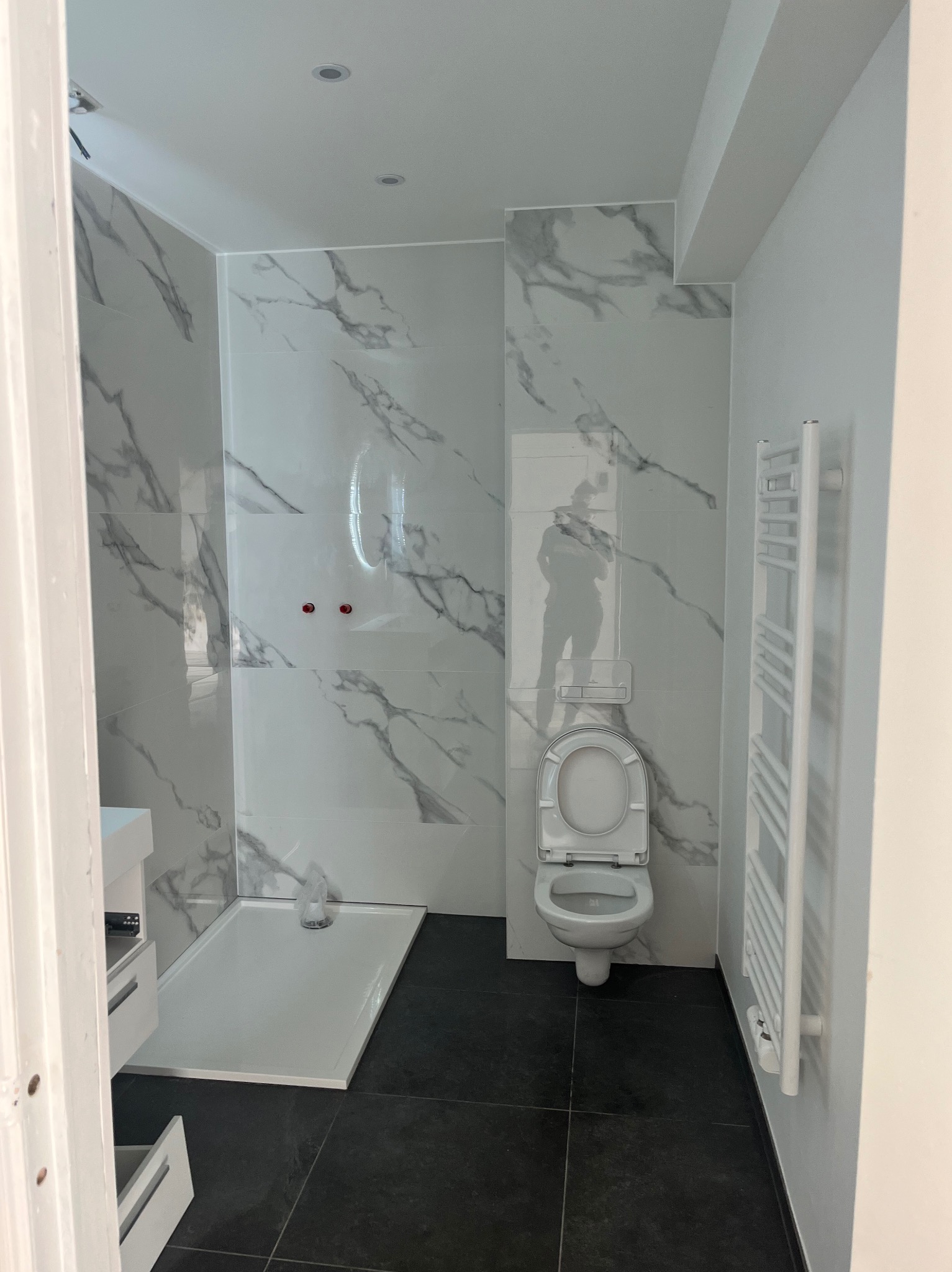 Wykończona łazienka z marmurowymi płytkami, czarną podłogą i białymi elementami: toaleta, grzejnik drabinkowy, brodzik. Widoczne czerwone zawory na ścianie.