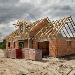 Profesjonalna budowa domów - Wznoszenie domu z czerwonej cegły: ściany, otwory okienne i dach w trakcie budowy, drewniana konstrukcja więźby dachowej, palety cegieł na pierwszym planie.