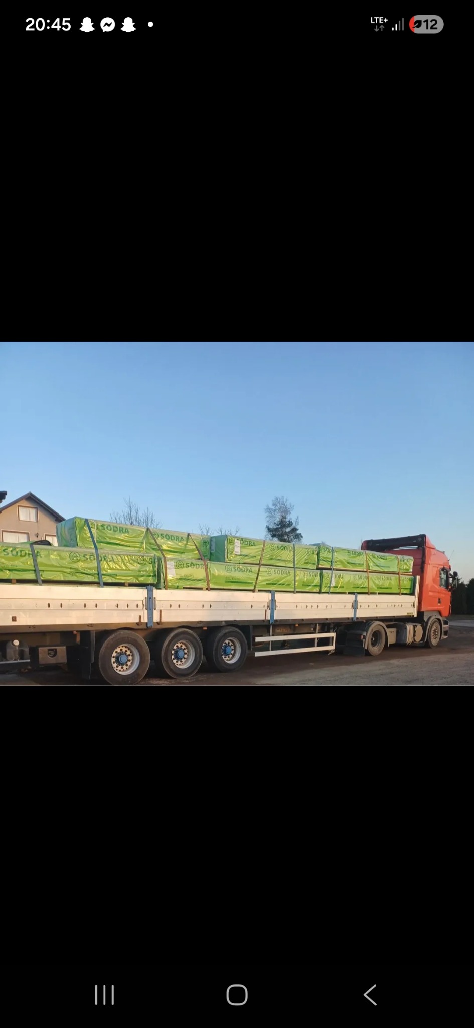 Ciężarówka z naczepą przewozi ładunek drewna budowlanego w zielonych opakowaniach marki Sodra, widok z boku na tle błękitnego nieba i zabudowań.