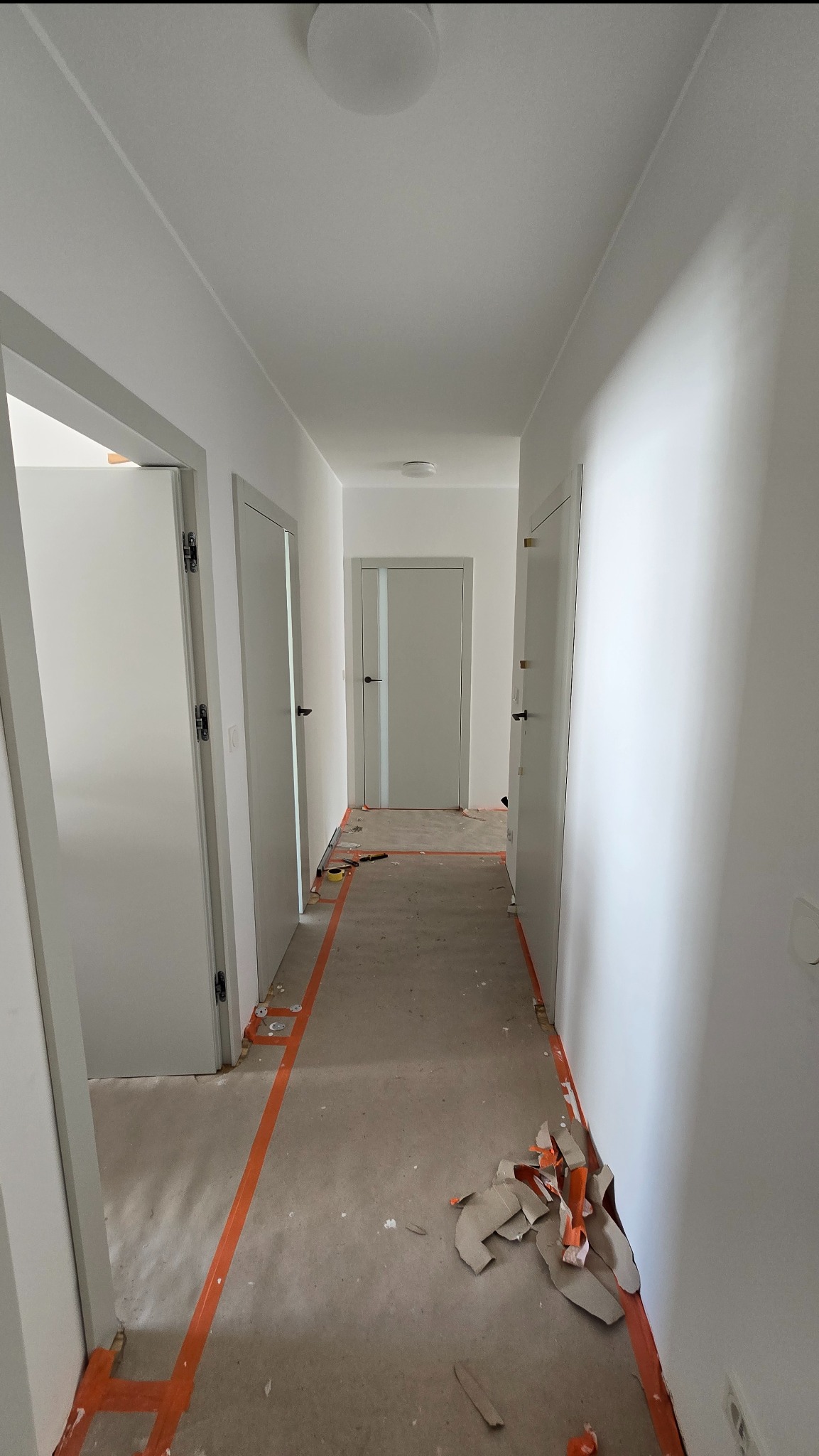Korytarz w trakcie montażu drzwi wewnętrznych z ościeżnicami w kolorze białym. Podłoga zabezpieczona taśmą malarską. Widoczne kartony po opakowaniach.