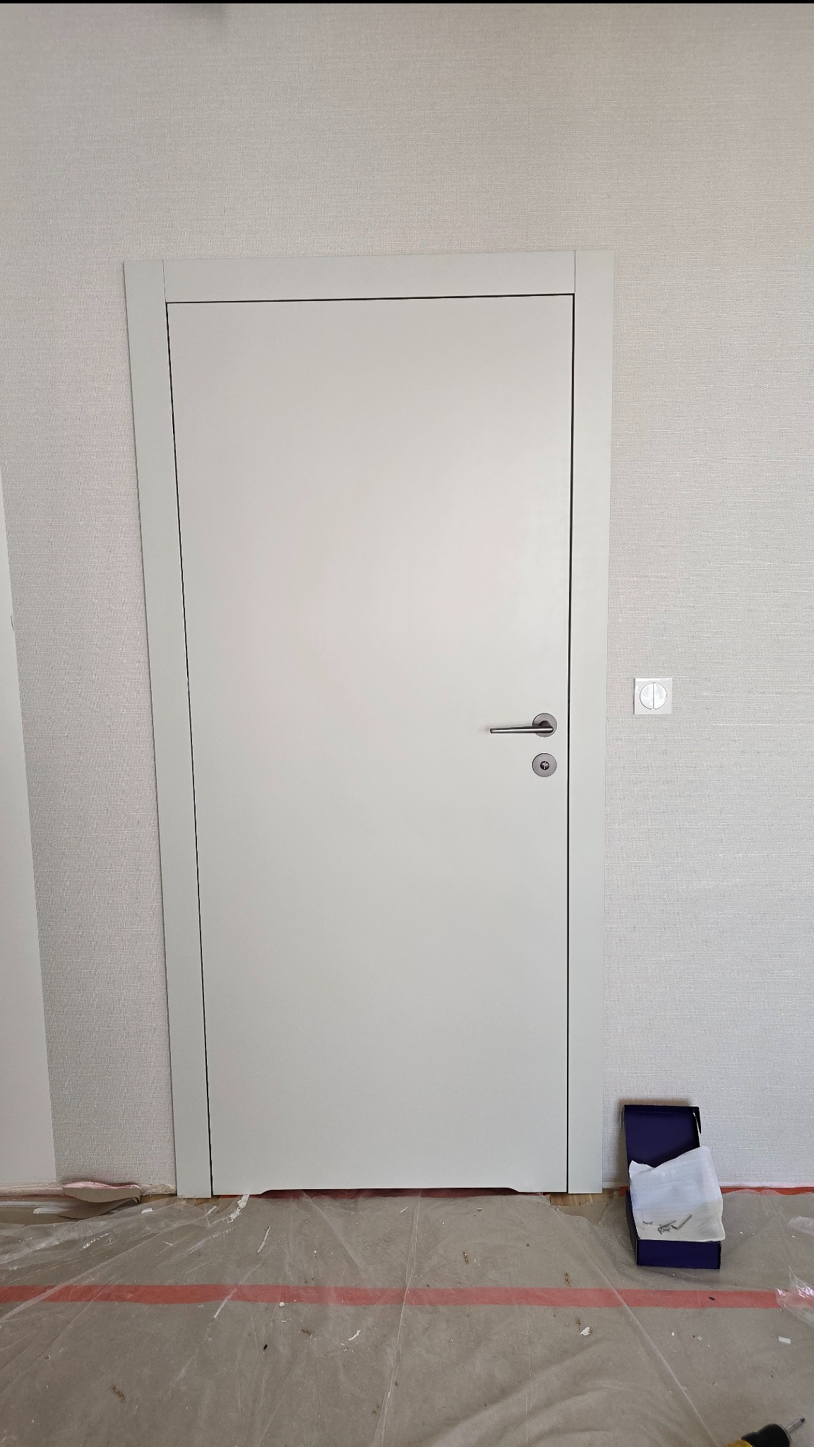 Białe, minimalistyczne drzwi wewnętrzne z srebrną klamką, zamontowane w pokoju z jasną tapetą. Na podłodze folia ochronna i pudełko z akcesoriami.