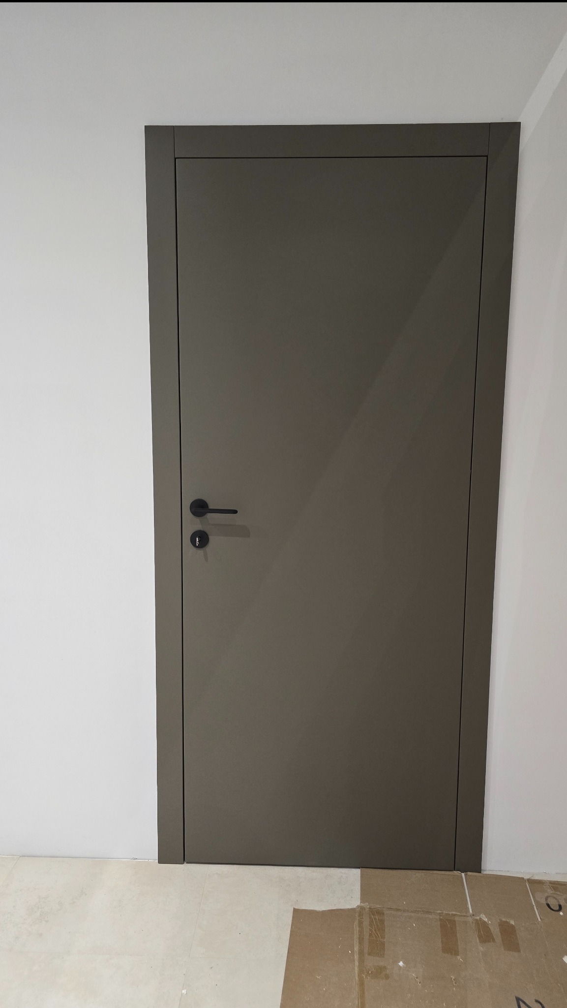 Zamknięte, gładkie, szare drzwi wewnętrzne z czarną klamką na białej ścianie. Widoczny fragment podłogi i kartonu. Minimalistyczny design.