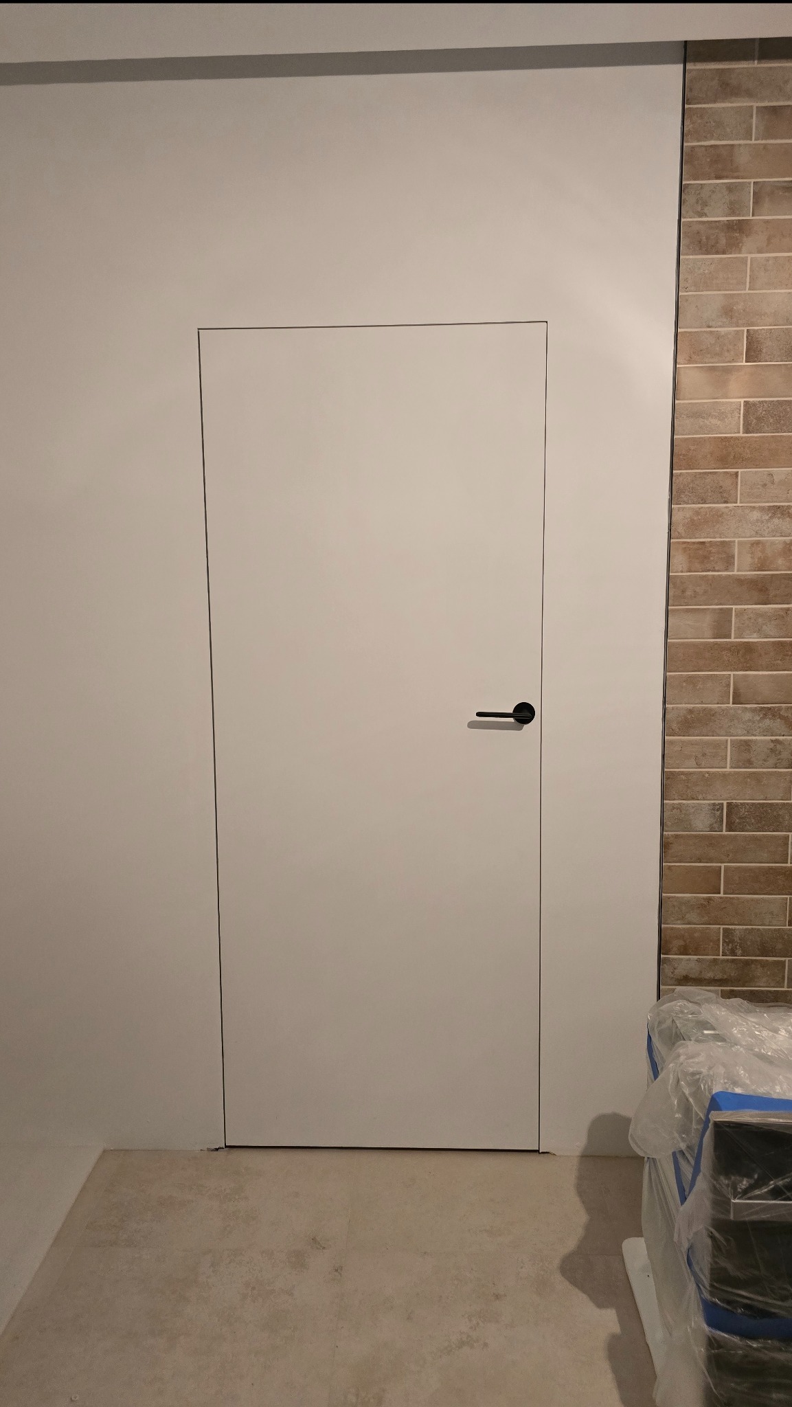 Białe, minimalistyczne drzwi z czarną klamką, wkomponowane w ścianę, obok fragmentu ściany wyłożonej cegłą. Widoczne materiały budowlane w folii.