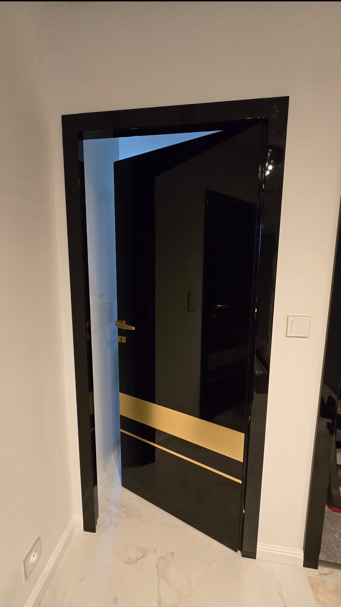 Częściowo otwarte, czarne drzwi wewnętrzne z połyskującą powierzchnią i złotymi pasami, widoczne na tle jasnej ściany i marmurowej podłogi.