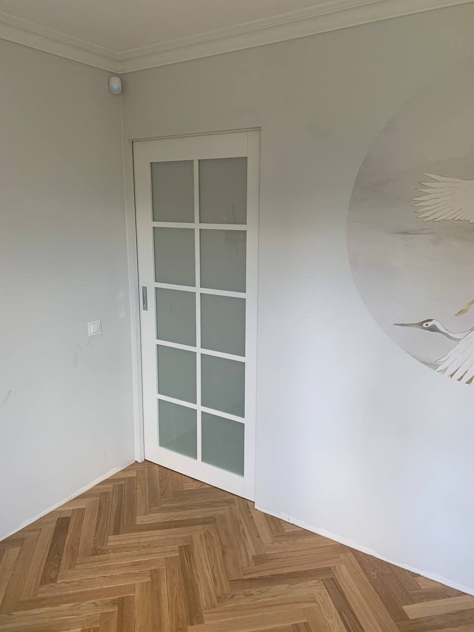 Białe, przeszklone drzwi przesuwne z podziałami na kwadraty, wkomponowane w jasne wnętrze z parkietem w jodełkę i malowidłem ptaka na ścianie.