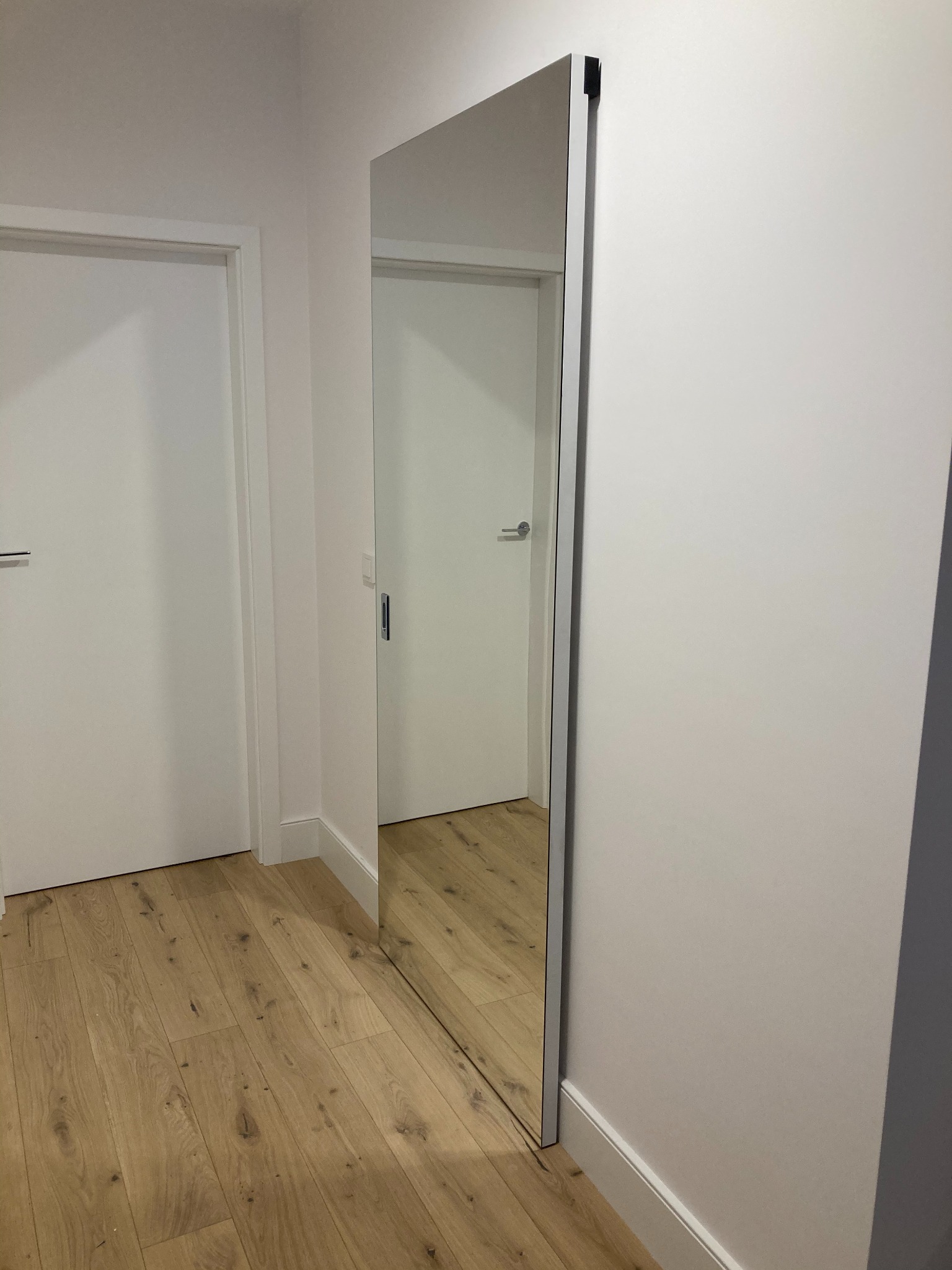 Minimalistyczne wnętrze z lustrzanymi drzwiami przesuwnymi w metalowej ramie, odbijającymi fragment drugiego pomieszczenia z białymi drzwiami i drewnianą podłogą.