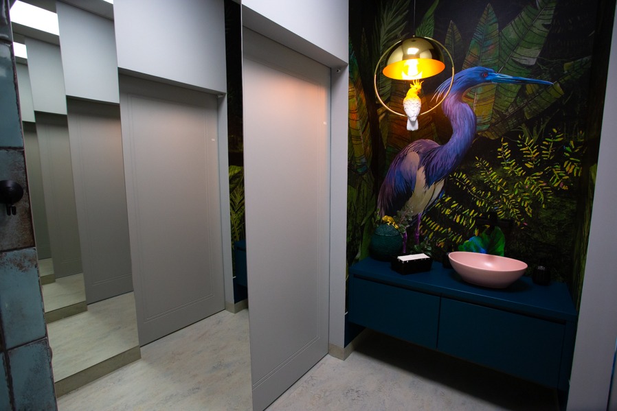 Szare drzwi wewnętrzne z lustrzanym wykończeniem, widoczne w rzędzie, z dekoracyjną tapetą z motywem tropikalnym i lampą w tle. Nietypowe wykończenie łazienki.
