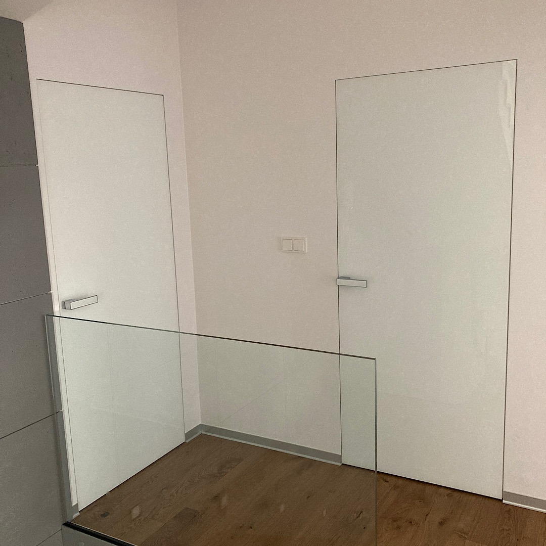 Dwie białe, minimalistyczne drzwi wewnętrzne z ukrytymi ościeżnicami i srebrnymi klamkami, widoczne za szklaną balustradą na tle jasnej ściany i drewnianej podłogi.