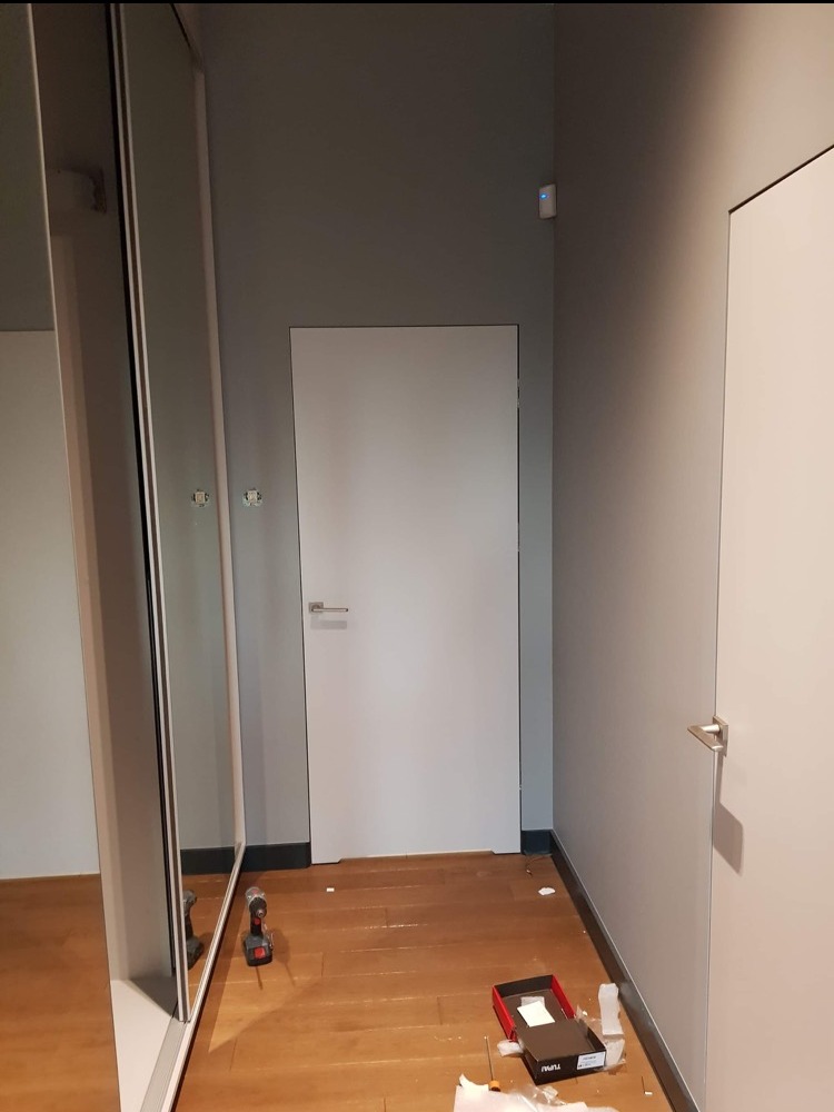 Montaż białych drzwi wewnętrznych z srebrną klamką. Na podłodze leżą narzędzia i akcesoria montażowe. Widoczna szafa z lustrem.