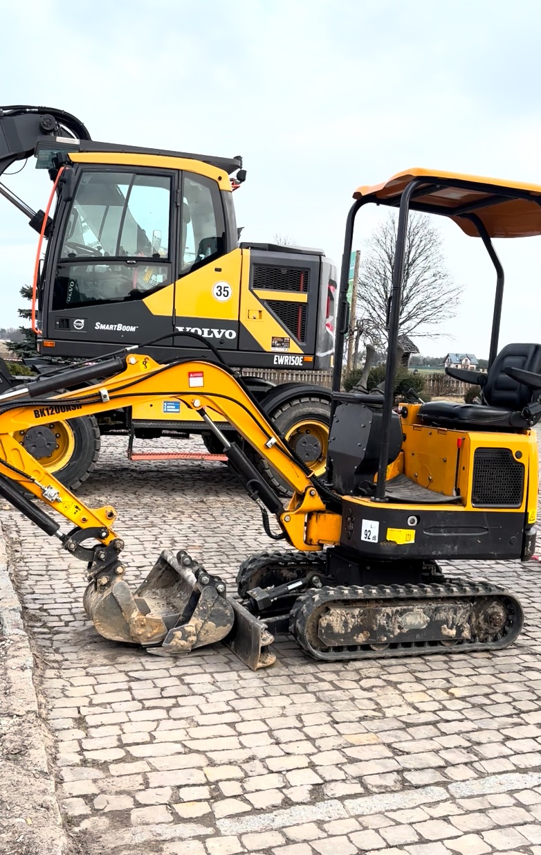 Żółta minikoparka Volvo BK1200KSH na brukowanej nawierzchni, z widoczną większą koparką w tle. Maszyna z wyraźnymi śladami użytkowania.