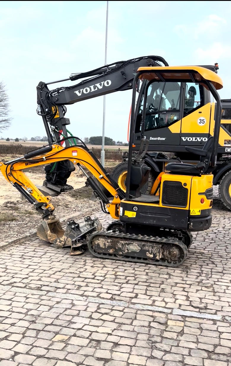 Żółto-czarna minikoparka Volvo na brukowanej nawierzchni, z widocznymi śladami użytkowania na gąsienicach i łyżce. W tle pole i częściowo widoczna inna maszyna.