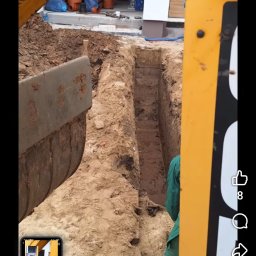 Artur-prace ziemne - Wąski wykop w ziemi, wykonany prawdopodobnie pod instalacje, widziany z perspektywy koparki. W tle widoczne elementy budynku i rury.