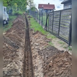 Artur-prace ziemne - Długi, wąski wykop w ziemi, prawdopodobnie pod instalacje, z widoczną ziemią po bokach i ogrodzeniem w tle. Widok z góry, perspektywa zbieżna.