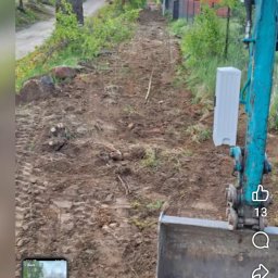 Artur-prace ziemne - Wąska droga gruntowa po niwelacji, widoczna z perspektywy operatora koparki. Ślady opon i wycięte pnie drzew. W tle zabudowania i zieleń.